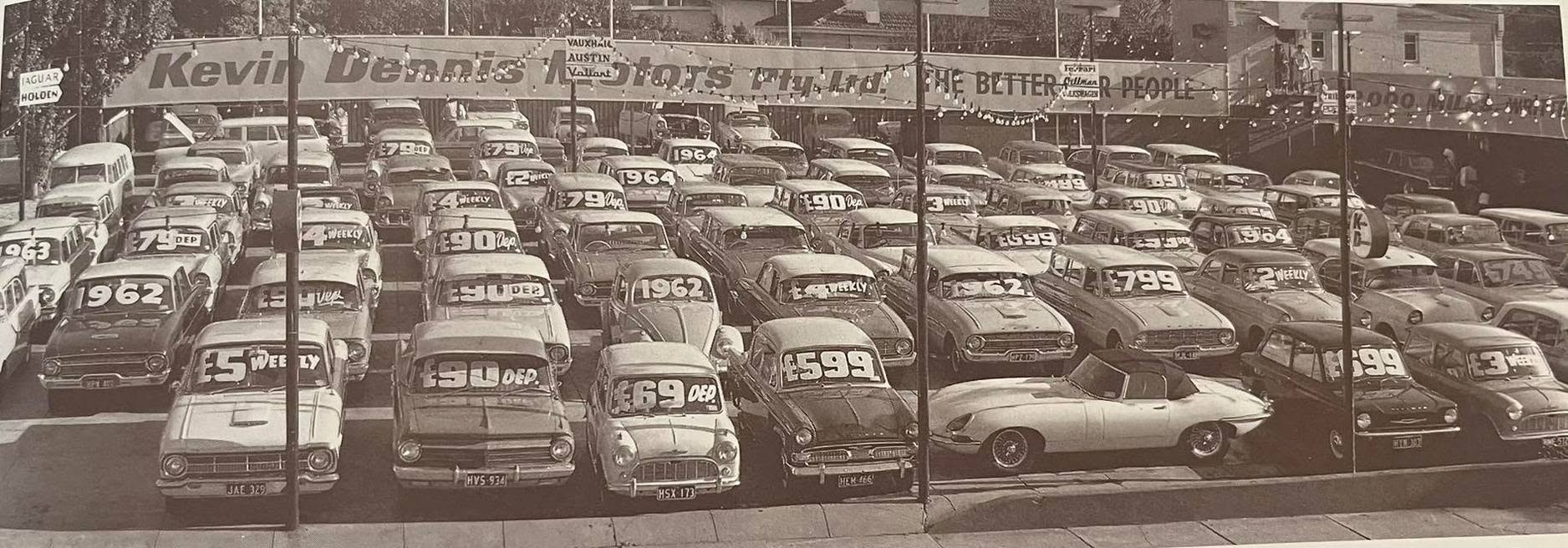 Vintage photograph of a packed Kevin Dennis Motors car yard displaying rows of Holden cars with large weekly-payment and deposit signs, with the dealership frontage and Holden branding visible above.